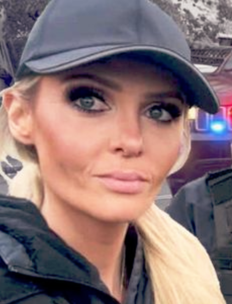 A photo of a heavily made up young woman wearing a black leather fascist cap and a puffy anorak, turning to look at the camera. Her hair, bound in a pony tail, has been draped over her left shoulder to ensure it enters the frame.
She is wearing heavy foundation, false eyelashes. Both her eyebrows, blusher and eye make-up all appear hastily applied. The head and shoulders shot with heavy pixelation around the edges has been overlaid rather amateurishly on a street scene.
 
The overall impression is of someone wishing to parody the kind of social media account that makes posts about being on the “front lines” of a made up confrontation.