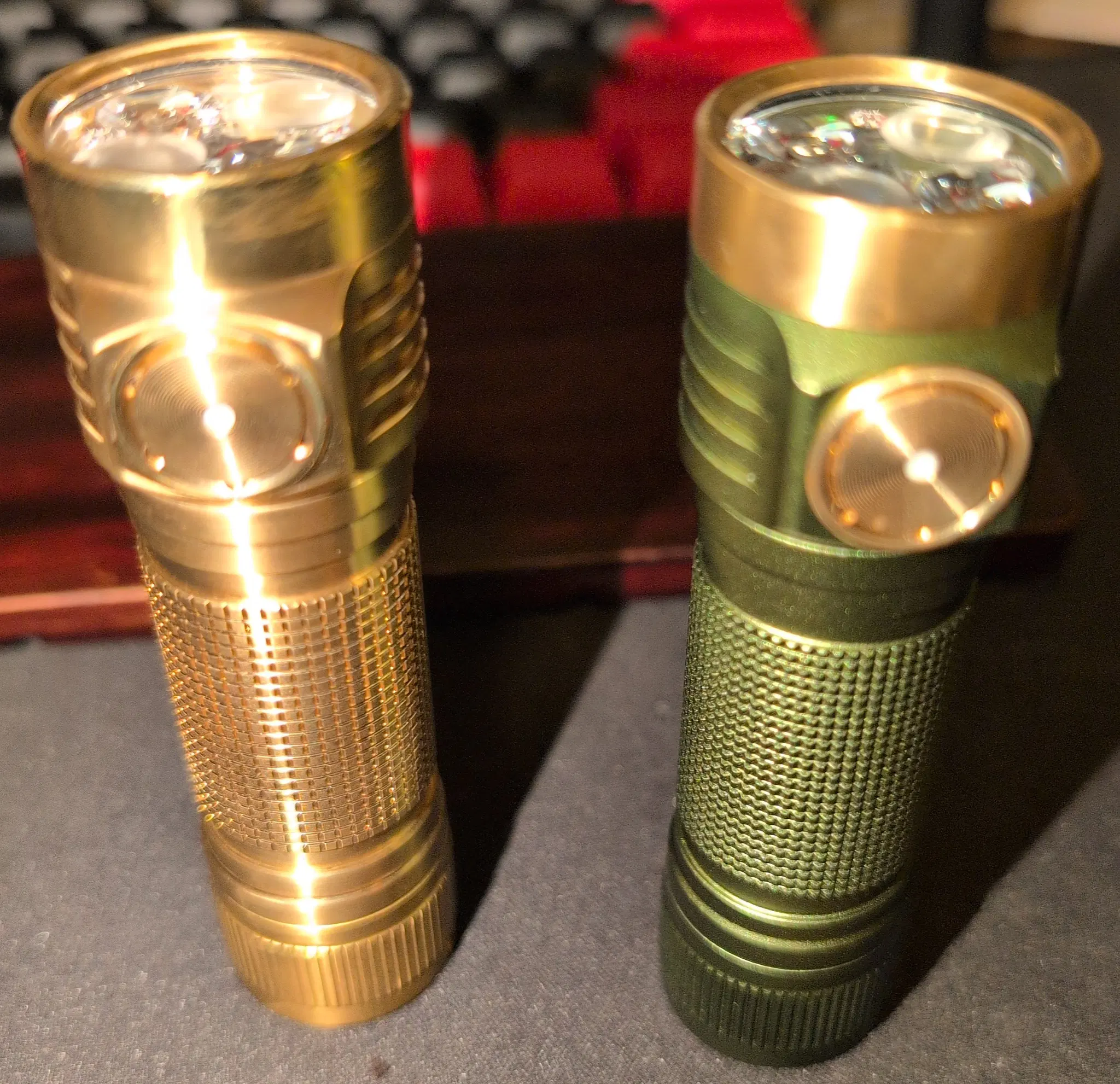 Two small metal LED flashlights standing upright. The left one is made of brass and the right one is green anodized aluminum.